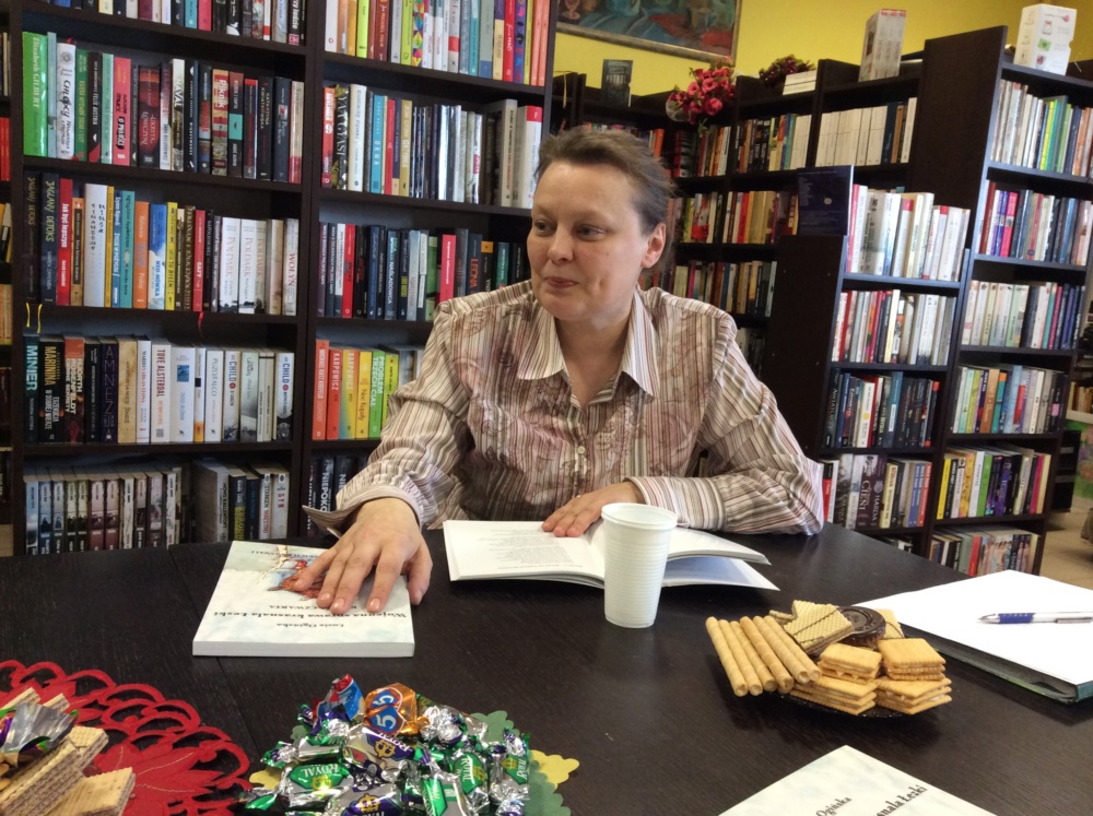 Światowy Dzień Poezji z Lusią Ogińską. Na zdjęciu autorka siedząca przy stole w BPGZ w Mokrem. Obok książki Pani Lusi, w tle regały biblioteczne.
