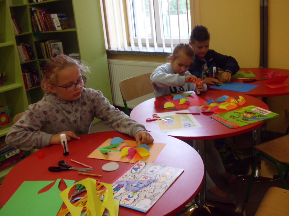 Wakacje 2019 Filia Żdanów. Na zdjęciu troje dzieci przy stolikach wyklejają kolorowe kwiaty.
