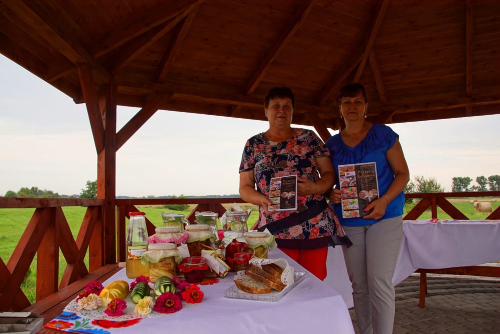 Premiera książeczki kucharskiej Pani Anny Zgnilec (na zdjęciu Pani Ania i Dyrektor Biblioteki Elżbieta Stankiewicz). Zdjęcie wykonane pod altanką. Panie stoją obok stołu zastawionego przetworami domowej roboty.